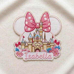 May include: An embroidered patch featuring a castle silhouette framed by mouse ears and a large pink polka-dot bow. The design includes blue and pink floral accents at the base and the name Isabella stitched in pink cursive lettering at the bottom.