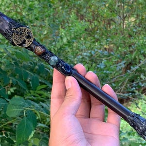 Op de afbeelding: Een donkere houten toverstaf met een puntige amethistkristal aan de bovenkant, een ronde goudkleurige levensboom-bedel en drie ingebedde gepolijste edelstenen in bruine, groene en blauwe tinten, met een textuurrijke basis en een rond uiteinde.