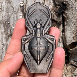 May include: A dark metallic, spider-shaped object with a smooth, polished finish. The spider features eight elongated legs and a central abdomen marked with a distinct, raised cross-like symbol. The overall design is symmetrical and flat.