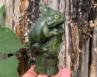 Puede incluir: Una escultura de piedra verde tallada que representa un camaleón posado sobre un tronco de árbol texturizado. El camaleón presenta una superficie lisa y pulida con una cresta distintiva en la cabeza y un ojo visible con iris amarillo.