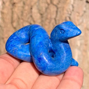 May include: A small, hand-carved snake figurine crafted from deep blue lapis lazuli stone. The sculpture features a coiled body with a smooth, polished surface, showing natural variations in the blue mineral texture and a defined head shape.
