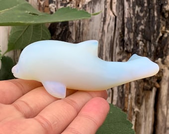 Puede incluir: Una figura de delfín lisa, translúcida y de color blanco lechoso, tallada en vidrio opalita. El delfín se muestra en posición de nado con una aleta dorsal curva, aletas pectorales laterales y una aleta caudal horizontal, con un suave brillo.