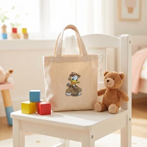 May include: A beige canvas tote bag featuring a central embroidered patch of a cartoon duck dressed in a tan explorer outfit with a pith helmet and a brown satchel strap across the chest. The bag has two matching fabric handles at the top.