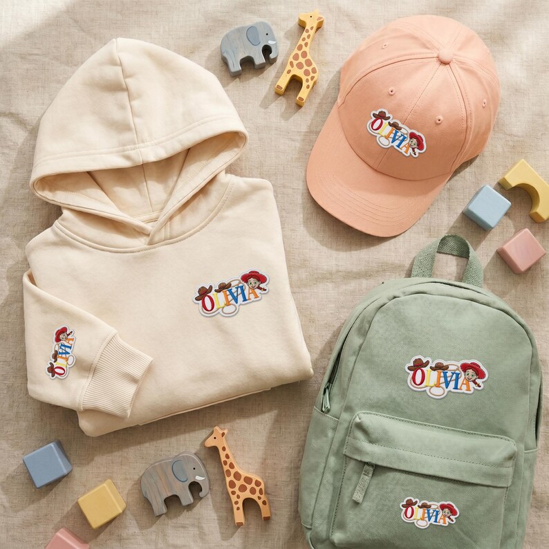 May include: A cream-colored hooded sweatshirt, a peach baseball cap, and a sage green backpack. Each item features an embroidered patch with the name Olivia in colorful letters, accented by cowboy hats and a cowgirl character graphic.