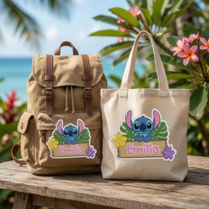 May include: A tan canvas backpack with brown leather straps and a matching off-white canvas tote bag. Both bags feature an embroidered patch of a blue alien character behind a wooden sign with the name Emilia, accented by green leaves and flowers.