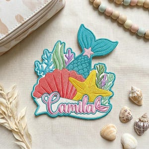 May include: An embroidered patch featuring a teal mermaid tail, a coral-colored seashell, and a yellow starfish surrounded by green and purple seaweed. A white banner across the bottom displays the name Camila in pink cursive script lettering.