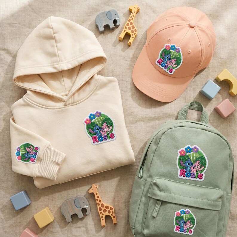 May include: A cream-colored hoodie, a peach baseball cap, and a sage green backpack, each featuring a circular patch with the characters Stitch and Angel and the name Rosie. Small wooden animal figures are scattered around the items.