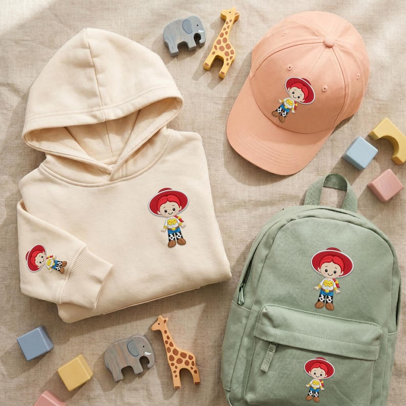 May include: A cream-colored hooded sweatshirt, a peach-colored baseball cap, and a sage green backpack, each featuring a patch of a cartoon cowgirl character with a red hat, yellow shirt, and blue pants, accompanied by small wooden animal figures.