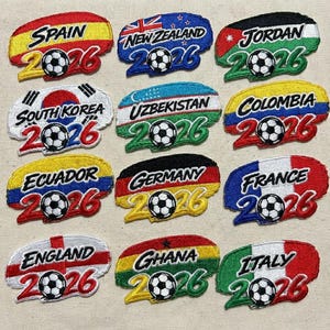 May include: Embroidered patches featuring flags of Spain, New Zealand, Jordan, South Korea, Uzbekistan, Colombia, Ecuador, Germany, France, England, Ghana, and Italy. Each patch includes a soccer ball and the year 2026.