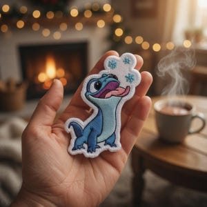 May include: Embroidered patch featuring a blue lizard with a pink tongue and snowflakes. The patch is held in a hand, with a warm beverage and fireplace in the background. The patch is a fun, decorative item.