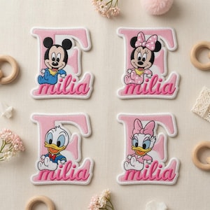 Könnte beinhalten: Vier gestickte Aufnäher mit dem Buchstaben E in Hellrosa und dem Namen Milia in Schreibschrift. Jeder Aufnäher zeigt eine andere Disney-Figur in einer Baby-Version: Mickey Maus, Minnie Maus, Donald Duck und Daisy Duck.