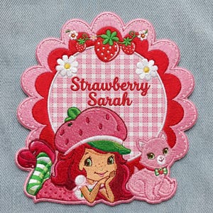 May include: Embroidered patch featuring a cartoon character with red hair and a strawberry hat, the text "Strawberry Sarah", and a pink cat. The patch has a scalloped edge with red and pink layers, and a gingham background.