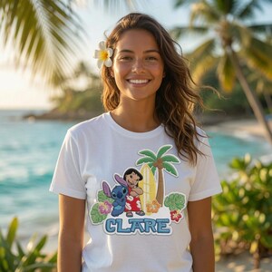 Peut inclure: Un t-shirt blanc &agrave; manches courtes avec une impression graphique des personnages de dessin anim&eacute; Lilo et Stitch &agrave; c&ocirc;t&eacute; d'une planche de surf jaune et d'un palmier, entour&eacute;s de fleurs tropicales et du nom CLARE en lettres bleues.