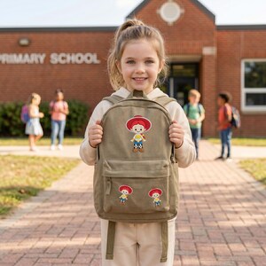 May include: A tan canvas backpack featuring three embroidered patches of a cartoon cowgirl character with red hair, a red hat, and a yellow shirt. The backpack has a large main compartment, a front zippered pocket, and adjustable shoulder straps.