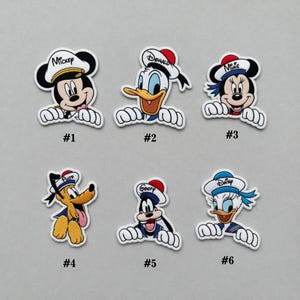 Peut inclure: Ensemble de six écussons de personnages Disney : Mickey Mouse, Donald Duck, Minnie Mouse, Pluto, Dingo et Daisy Duck. Chaque écusson a une bordure blanche et un nom de personnage sur le chapeau.