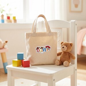 May include: A beige canvas tote bag with two handles featuring a colorful graphic of the name OLIVIA in block letters, decorated with a cowboy hat, a lasso, and a cowgirl character illustration centered on the front of the fabric bag.