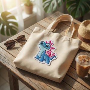 May include: A beige canvas tote bag with a blue and pink cartoon character patch. The bag sits on a wooden table next to sunglasses, a straw hat, and a cold beverage in a clear plastic cup.