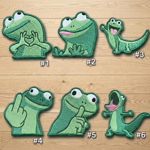 May include: A collection of six embroidered patches featuring cartoon frogs and lizards in various poses. The patches are primarily green with white and black eyes, and pink accents. Each patch has a number from #1 to #6.
