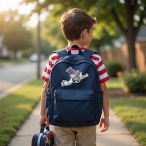 May include: A navy blue backpack with a Buzz Lightyear graphic, carried by a child. The backpack has a front pocket and side pockets. The child is wearing a red and white striped shirt and khaki pants, carrying a matching lunch bag.