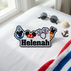 以下が含まれることがあります： 黒い文字で「Helenah」と書かれた白い刺繍ワッペン。テキストの上には、ミッキーの耳が付いた青い錨、赤と黄色のミッキーの頭、青と白のストライプのヘッドバンド、ミッキーのアイスクリームという4つのアイコンがデザインされています。