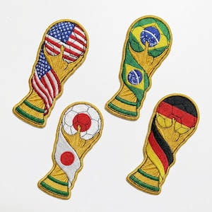 Puede incluir: Parches bordados con el diseño del trofeo de la Copa del Mundo, cada uno con una bandera diferente: EE. UU., Brasil, Japón y Alemania. Los parches tienen detalles dorados y verdes, para ropa o accesorios.