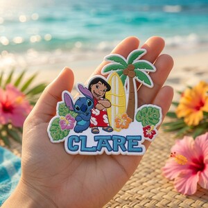 Peut inclure: Un &eacute;cusson brod&eacute; repr&eacute;sentant les personnages Lilo et Stitch &agrave; c&ocirc;t&eacute; d'une planche de surf jaune et d'un palmier, avec le nom CLARE en lettres bleues en bas, entour&eacute; de feuilles tropicales vertes et de fleurs d'hibiscus color&eacute;es.
