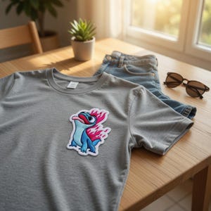 May include: A gray t-shirt with a colorful patch of a cartoon salamander with blue, red, and pink flames. The shirt is paired with blue jeans and sunglasses on a wooden table, suggesting casual wear.