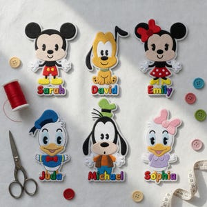 May include: Six embroidered character patches featuring Mickey, Pluto, Minnie, Donald, Goofy, and Daisy. Each patch includes a name in colourful lettering: Sarah, David, Emily, Juda, Michael, and Sophia. Includes red thread, scissors, and buttons.