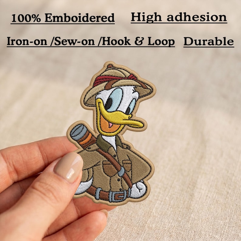 May include: An embroidered patch featuring a cartoon duck dressed as an explorer in a tan jacket and hat with a red band. Text above reads 100% Emboidered, High adhesion, Iron-on /Sew-on /Hook & Loop, and Durable. The patch has a tan border.
