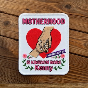 May include: Embroidered patch featuring the word "MOTHERHOOD" in pink above a red heart and clasped hands. The text "IS KINGDOM WORK Kenny" is below the heart, with a purple banner reading "PROVERBS 31:25-27". Floral accents surround the text.