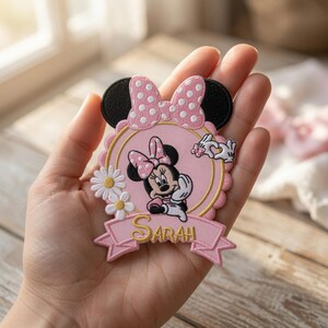 Puede incluir: Un parche bordado que presenta un marco circular rosa con borde festoneado, el personaje de Minnie Mouse, un lazo rosa con lunares, dos margaritas blancas y una cinta rosa en la parte inferior con el nombre Sarah bordado en hilo dorado.