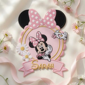 Puede incluir: Un parche bordado decorativo que presenta un rat&oacute;n de dibujos animados con un lazo rosa de lunares, dentro de un marco rosa festoneado. El dise&ntilde;o incluye margaritas blancas, un par de manos con guantes blancos formando un coraz&oacute;n y una cinta rosa con el nombre Sarah.