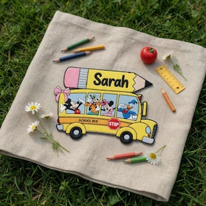 Può includere: Un sottopentola in lino beige con una toppa a forma di scuolabus giallo con personaggi Disney e il nome "Sarah". La scena comprende matite, una mela rossa, un righello e margherite.