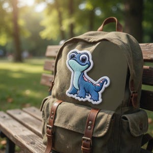 May include: A blue and white embroidered patch of a cartoon lizard is affixed to a green canvas backpack. The backpack has brown leather straps and buckles. The patch features a light blue lizard with pink accents.