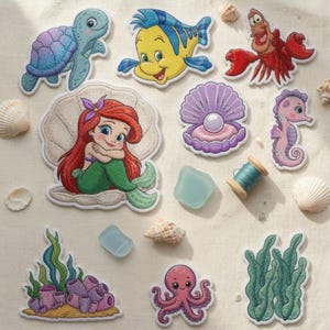 May include: A collection of embroidered fabric patches featuring a mermaid, a turtle, a yellow fish, a red crab, a seahorse, an octopus, a pearl in a shell, and seaweed, accompanied by a small spool of blue thread and scattered seashells.