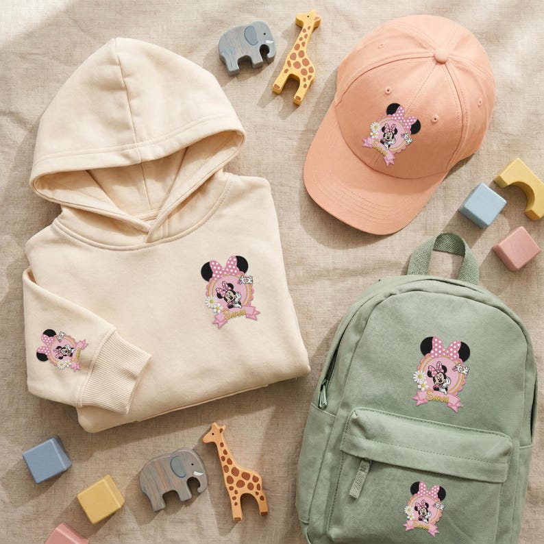 Puede incluir: Una sudadera con capucha beige, una gorra de b&eacute;isbol color melocot&oacute;n y una mochila verde salvia, cada una con un gr&aacute;fico de Minnie Mouse con el texto "Sweet". Los art&iacute;culos est&aacute;n dispuestos con figuras de animales de madera y bloques.