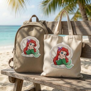 May include: A tan canvas backpack and a matching off-white canvas tote bag, both featuring a centred embroidered patch of a cartoon mermaid with long red hair and a green tail, sitting inside a large white seashell with a purple hair accessory.