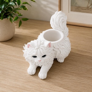 Puede incluir: Una maceta con forma de gato blanco que presenta una textura de pelo largo y esponjoso con un esculpido detallado del pelaje. El gato está en posición agachada con la cola curvada hacia arriba y una abertura cilíndrica integrada en el lomo.