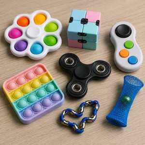 May include: A collection of colorful fidget toys, including a flower-shaped popper, a cube, a spinner, a controller-style toy, a rainbow popper, a blue mesh tube with a marble, and a blue and silver linked chain. The toys are on a light wood surface.