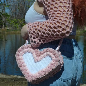 May include: A heart-shaped, pink and white crochet bag with a matching pink crochet sleeve. The bag has a white center and a pink border. The sleeve is a light pink, open weave design. The bag is held in front of a body wearing blue jeans.