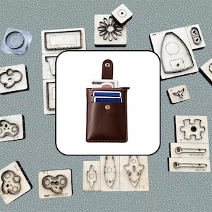 May include: A dark brown leather card holder with a vertical orientation, featuring a snap-button closure strap, multiple exterior card slots, and a side zipper. The card holder is surrounded by various metal cutting dies on a textured gray surface.