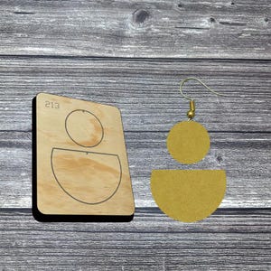 May include: A wooden earring die with the number "213" and a pair of mustard yellow suede earrings. The earrings feature a circular top and a semi-circular bottom. The die is made of light-colored wood.