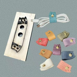 May include: A collection of cat-shaped cable organizers in various colors, including blue, pink, and purple. The organizers are made of leather and feature a gold snap closure. A wooden cat-shaped template is also present.