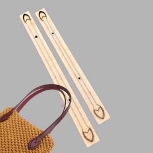 Può includere: Modelli in legno per manici di borse con design a forma di cuore, accanto a una borsa intrecciata con manico in pelle bordeaux. I modelli sono marrone chiaro con dettagli incisi e la borsa è color marrone dorato.