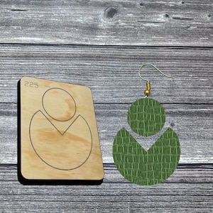 May include: A pair of green leather earrings with a textured, crocodile-like pattern. The earrings are in a geometric design, with a circular top and a crescent shape below. A wooden die with the number 225 and the earring shape is next to the earrings.