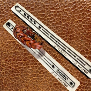 May include: A brown leather braided bracelet with a metal buckle rests on a white template. The template has the text "2025.12.28" and other text. The background is a textured brown surface.