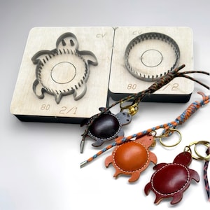 May include: Two wooden cutting dies with metal blades, one shaped like a turtle and one circular, both marked 80 2/1. Three leather turtle keychains in dark brown, orange, and red, each attached to a multicolored braided cord and gold ring.
