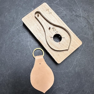 May include: A light brown leather keychain with a brass ring next to a wooden die-cut tool. The die-cut tool has a teardrop shape with the text "2-2" and "Ø2.0" printed on it. The keychain is made of a soft, textured leather.