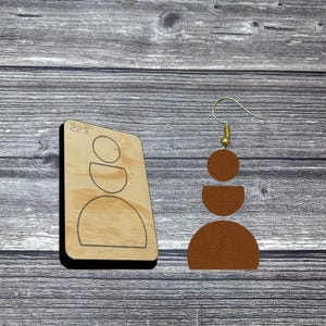 May include: A pair of brown leather earrings with a gold hook, featuring a geometric design of stacked circles and a half-circle. A wooden die-cut with the number "223" is also present.