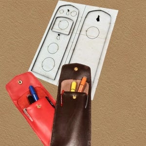 May include: A red and a brown leather pen case, each with a snap closure, holding pens and markers. A paper template with a design for the pen cases is in the background. The pen cases are on a tan surface.